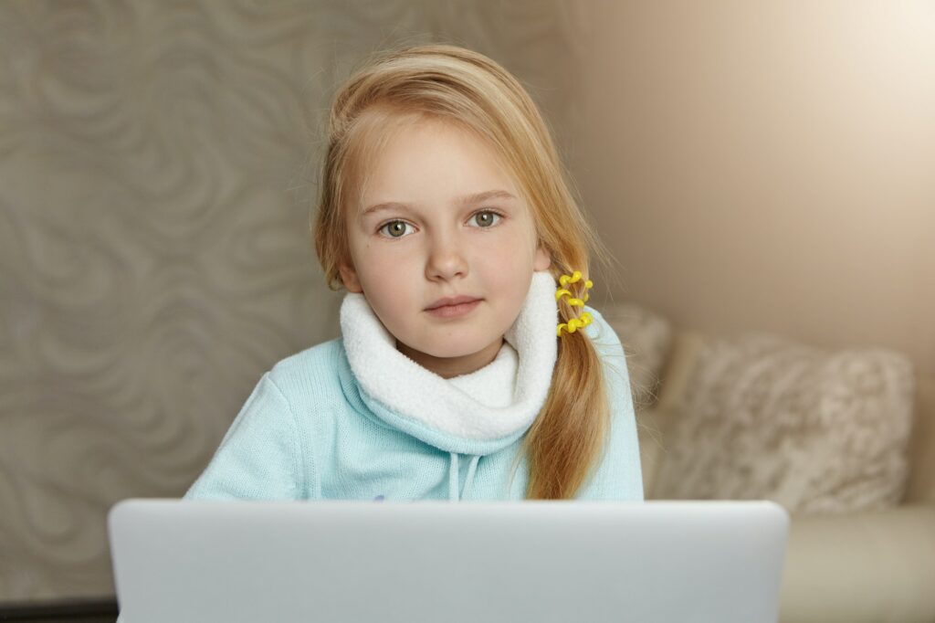 Adorable blonde female school kid with messy ponytail enjoying leisure time at home, sitting in her